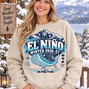 May include: A beige crewneck sweatshirt featuring a graphic print with the text EL NINO WINTER 2026-27. The design includes a map of the United States with weather patterns, snowflakes, clouds, and text reading COLDER, WETTER, STORMIER and A STRONGER WINTER RETURNS.