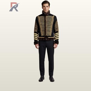 Op de afbeelding: Zwarte jas in militaire stijl met gouden details en een bontkraag. De jas heeft gouden vlechtwerk en borduursels op de mouwen. De persoon draagt een zwarte broek en schoenen.