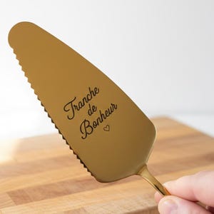 Könnte beinhalten: Ein goldfarbener Tortenheber mit gezahntem Rand und einem herzförmigen Detail. Der Heber trägt die Worte "Tranche de Bonheur" in Schreibschrift. Der Heber wird über einem Holzschneidebrett gehalten.
