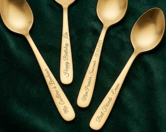Puede incluir: Cuatro cucharas de metal en tono dorado con texto grabado en los mangos. Las inscripciones dicen: Ma Cuillère à Bonheur, Happy Birthday - Léo, Mon Premier Souvenir - 12.03.2025 y Best Friends - Emma. Las cucharas están dispuestas sobre tela verde.