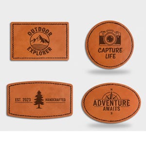 Peut inclure: Quatre écussons en cuir marron avec des bords cousus noirs. Les écussons présentent des motifs sur le thème du plein air et du voyage, notamment une scène de montagne, un appareil photo, un arbre et une boussole avec les mots "Outdoor Explorer", "Capture Life", "Handcrafted" et "Adventure Awaits".