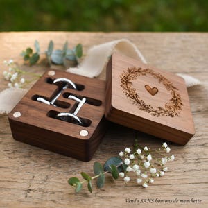 May include: A dark brown wooden box with a lid featuring a laser-engraved floral wreath and a central heart. The open box contains two silver-toned metal cufflinks. The box is square with rounded corners and visible magnetic closures.