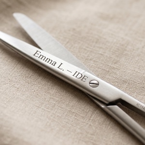 May include: Close-up of stainless steel scissors with the text "Emma L. - IDE" engraved on the handle. The blades are open, revealing a sharp edge. The scissors rest on a neutral-toned fabric surface.