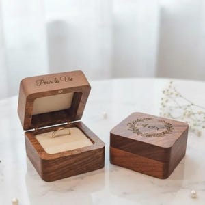 Pode incluir: Duas caixas de anel de madeira. Uma caixa está aberta, revelando um anel de ouro em uma almofada creme. A caixa aberta tem o texto "Pour la Vie" gravado na tampa. A caixa fechada tem um design de coroa com iniciais.