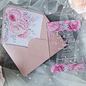 Può includere: Un set di inviti rosa blush con un invito in acrilico trasparente e una busta abbinata. L'invito e l'interno della busta sono adornati da un motivo floreale nei toni del rosa e del bianco. Il testo dell'invito recita "Lily's 70th Birthday".