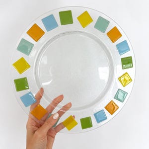 May include: A clear glass plate featuring a circular border of small, square-shaped glass tiles in various colours including light blue, lime green, yellow, and orange, arranged in a repeating pattern around the outer rim of the circular dish.