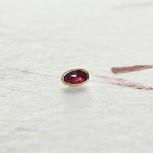 Könnte beinhalten: Ein kleiner, oval geformter Edelstein in einem Goldrahmen. Der Edelstein hat eine tiefrote Farbe und eine polierte Oberfläche, die das Licht reflektiert. Die Goldeinfassung bildet einen warmen Kontrast zum roten Stein.