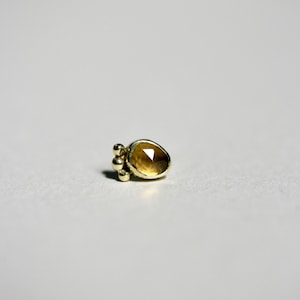 Pode incluir: Um pequeno brinco de pressão dourado com uma pedra preciosa âmbar facetada. Três pequenas contas douradas estão agrupadas à esquerda da pedra preciosa. O brinco está sobre um fundo branco liso.