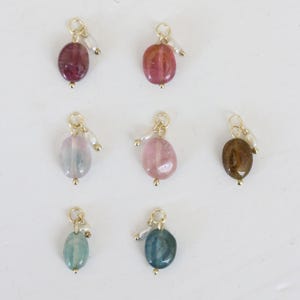 Könnte beinhalten: Sieben Edelsteinanhänger in verschiedenen Farben, darunter Rosa, Blau und Braun. Jeder Anhänger hat eine goldfarbene Schlaufe und einen kleinen Perlenakzent. Die Edelsteine sind oval und poliert.