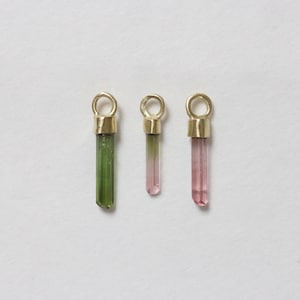 Op de afbeelding: Drie edelsteenhangers met goudkleurige doppen en lussen. De edelstenen zijn cilindrisch, met één groene, één roze en één roze-groene. De hangers worden gepresenteerd tegen een witte achtergrond.