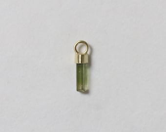 May include: A small, green tourmaline crystal pendant with a gold-colored bail. The tourmaline is a translucent, light green color and is rectangular in shape. The bail is a simple, circular design.