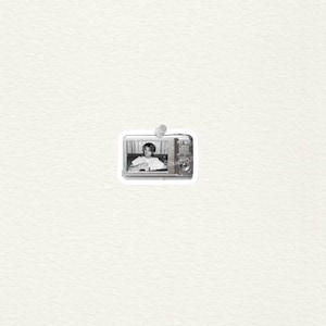 May include: Monochrome sticker depicting a vintage camera with a photograph of a person inside. The sticker is affixed to a white surface with a drawing pin. The camera has a retro design with dials and a lens.