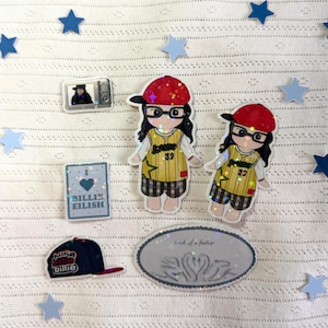 May include: A collection of stickers and a baseball cap featuring a cartoon girl wearing glasses and a red hat. The stickers include a small TV, a heart-shaped design with the text "Billie Eilish", and an oval sticker with the text "birds of a feather".