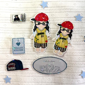 May include: A collection of stickers and pins featuring a cartoon character wearing a red baseball cap and glasses, a blue baseball cap, a television, and the text "Billie Eilish". The stickers are surrounded by blue stars.