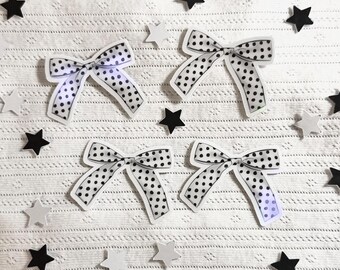 May include: Four white bows with black polka dots are arranged on a white textured surface, interspersed with black and white star-shaped decorations. The bows have a classic shape with a contrasting black and white pattern.