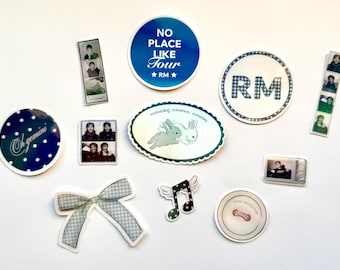 May include: A collection of stickers with various designs. Includes a blue circle sticker with the text "NO PLACE LIKE TOUR" and "RM" stars, a sticker with the letters "RM" in a checkered pattern, and stickers with photo booth style images.