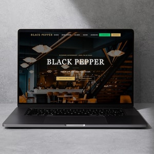 以下が含まれることがあります： Black Pepperレストランのウェブサイトを表示しているラップトップ。ウェブサイトには、レストランの暗いインテリアの写真と、白い文字で「Black Pepper」という言葉が表示されています。ラップトップは、明るい灰色の表面に開いています。