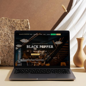 Può includere: Un laptop mostra il sito web "BLACK PEPPER" con un interno di ristorante. Il laptop è su una superficie beige, accanto a un blocco di pietra, due vasi di ceramica e un tessuto bianco. Il sito web ha uno schema di colori scuri.