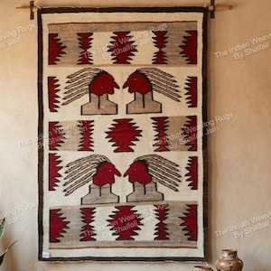 Puede incluir: Un tapiz tejido con un diseño nativo americano. La alfombra presenta un fondo crema con patrones geométricos rojos y grises y dos figuras humanas estilizadas con caras rojas y tocados de plumas. La alfombra se exhibe en un pasador de madera.
