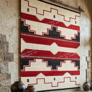 Puede incluir: Una alfombra mural rectangular grande con un patrón geométrico en rojo, negro y crema. La alfombra está suspendida de una barra decorativa de hierro forjado. El diseño presenta líneas escalonadas y bandas horizontales, creando una estética del suroeste.