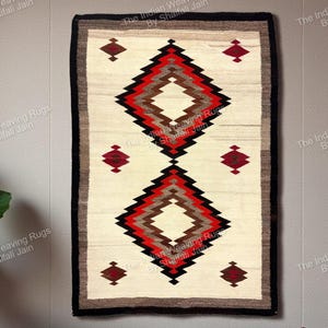 Op de afbeelding: Een rechthoekige geweven wandhanger met een crèmekleurige achtergrond en een geometrisch ontwerp in rood, bruin en zwart. Het ontwerp heeft twee ruitvormen en kleinere ruitaccenten. De rand is zwart en bruin.