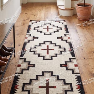 May include: Cream-colored runner rug with a geometric pattern in black, gray, and red. The rug features a central cross design and is placed on a wooden floor in a hallway. The rug is labeled "SHAILI JAIN".