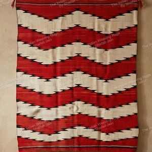 Op de afbeelding: Een rood, wit en zwart geometrisch patroon tapijt dat aan een muur hangt. Het tapijt heeft een zigzagontwerp met afwisselende banden van rood en wit, geaccentueerd met zwarte ruitvormen. Kwasten hangen aan de boven- en onderranden.