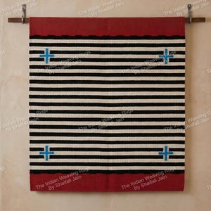Pode incluir: Um tapete retangular tecido com listras horizontais pretas e brancas, bordas vermelhas e quatro desenhos de cruzes azuis. O tapete está pendurado em uma haste de madeira contra uma parede bege. O texto "The Indian Weaving Rugs By Shaifali Jain" é visível.