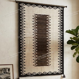 May include: A rectangular wall hanging rug with a geometric design. The rug features a cream-colored center with a dark brown rectangular accent and a black border with diamond-shaped patterns. It is mounted on a dark brown rod.