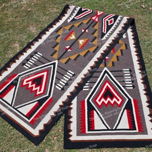 Puede incluir: Una alfombra rectangular tejida a mano con un patrón geométrico. La alfombra presenta un diseño central en tonos grises, negros, rojos, blancos y beige. El diseño incluye flechas, diamantes y formas en zigzag. La alfombra probablemente está hecha de lana o un material similar.