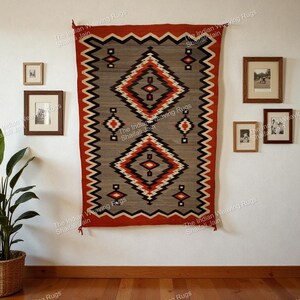 May include: A gray, red, and black geometric patterned rug hanging on a white wall. The rug features diamond and rectangular shapes. The rug is framed by a red border. The wall also has framed pictures and a potted plant.
