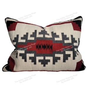 Puede incluir: Cojín rectangular con un patrón geométrico en crema, rojo, negro y gris. El diseño presenta formas repetidas y una sección central roja. El cojín tiene un aspecto suave y texturizado, adecuado para la decoración del hogar.