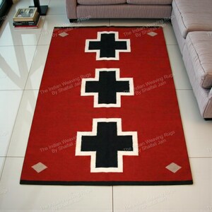 Pode incluir: Um tapete retangular vermelho com três grandes cruzes pretas delineadas em branco. O tapete tem uma borda preta e formas de diamante nos cantos. O texto "The Indian Weaving Rugs By Shaifali Jain" está impresso no tapete.