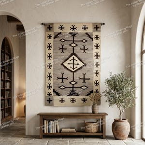 Pode incluir: Uma tapeçaria de parede tecida com um design geométrico em tons de cinza, creme e preto. O tapete é retangular e apresenta uma forma de diamante central com padrões de cruzes. Pendurado acima de um banco de madeira com livros e um vaso.