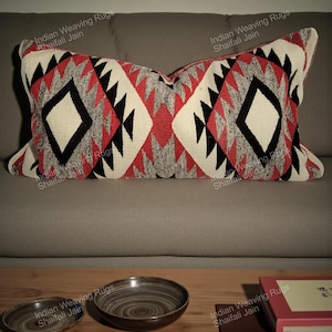 May include: A rectangular cushion with a geometric pattern in red, black, white, and grey. The cushion is on a grey sofa. The design features diamond and triangle shapes. The text "Indian Weaving Rugs Shaifali Jain" is visible on the cushion.