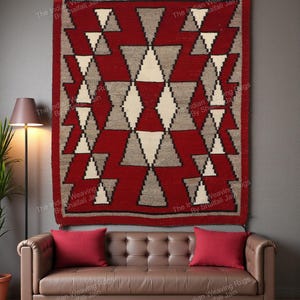 Può includere: Un tappeto rosso e grigio con motivi geometrici appeso a una parete. Il tappeto presenta forme a diamante e triangolo in rosso, grigio e bianco. Il tappeto è sopra un divano in pelle marrone con cuscini rossi.