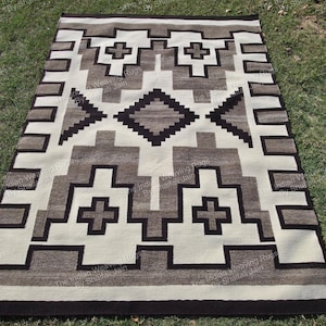 Pode incluir: Tapete retangular tecido com um padrão geométrico em tons de castanho, creme e preto. O design apresenta formas de cruz e diamante repetidas. O tapete é exibido sobre relva verde.