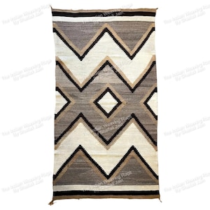 Può includere: Un tappeto rettangolare tessuto con un motivo geometrico. Il tappeto presenta una forma a diamante centrale e motivi a zig-zag nei toni del marrone, crema e nero. Il tappeto è realizzato in un materiale spesso e strutturato, con nappe alle estremità.
