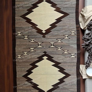 Pode incluir: Tapete retangular tecido com um design geométrico. O tapete apresenta um padrão central de diamante em bege e castanho, emoldurado por uma borda castanha escura. O fundo é uma mistura de tons cinza e castanho, criando uma aparência texturizada. O tapete é provavelmente feito de fibras naturais.