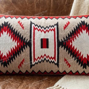 May include: A rectangular throw pillow featuring a beige base with a geometric pattern in red, black, and white. The design includes two large diamond shapes flanking a central rectangular motif, all bordered by a red zigzag edge trim.