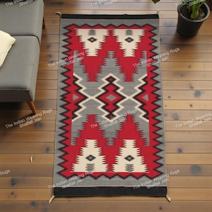 Può includere: Un tappeto rettangolare con un motivo geometrico rosso, grigio, nero e bianco. Il tappeto presenta un disegno centrale con forme a diamante e triangolo, incorniciato da un bordo a zig-zag. Il tappeto è posto su un pavimento in legno.