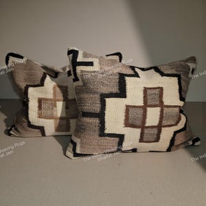 Puede incluir: Dos cojines decorativos con patrones geométricos. Los cojines tienen un fondo crema con formas geométricas marrones, negras y grises. Los cojines están hechos de tela tejida y están colocados sobre una superficie neutra.
