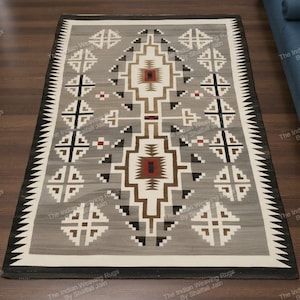 Può includere: Tappeto rettangolare grigio con un motivo geometrico marrone, nero e bianco. Il tappeto presenta un disegno centrale con forme ripetute e un bordo a zig-zag. È posato su un pavimento in legno. Misura circa 150 cm x 200 cm.