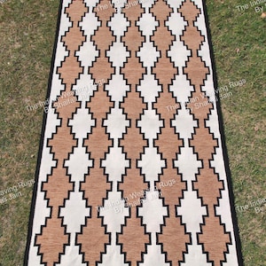 Puede incluir: Alfombra rectangular con un patrón geométrico de diamantes en marrón, blanco y negro. La alfombra está hecha de material tejido y se exhibe sobre una superficie cubierta de hierba. El diseño presenta formas de diamantes repetidas.