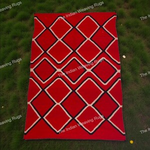 Op de afbeelding: Een rechthoekig rood tapijt met een geometrisch ruitpatroon in wit en zwart. Het tapijt is gemaakt van geweven materiaal en wordt getoond op een grasachtige ondergrond. De tekst "The Indian Weaving Rugs" staat op het tapijt.