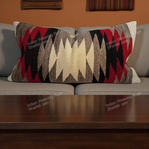 Puede incluir: Cojín decorativo rectangular con un patrón geométrico en rojo, negro, gris y crema. El cojín está sobre un sofá gris. El texto "Indian Weaving Rugs Shaifali Jain" está impreso en el cojín.