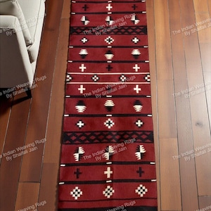 Puede incluir: Una alfombra rectangular de color burdeos con un patrón geométrico. El diseño incluye cruces blancas, formas de diamante y otros elementos geométricos, con rayas negras en la parte superior e inferior. La alfombra está sobre un suelo de madera.