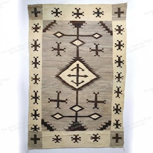 Può includere: Tappeto tessuto a mano con un motivo geometrico. Il tappeto presenta una forma a diamante centrale con simboli a forma di croce e un bordo di croci. La tavolozza dei colori comprende beige, grigio e marrone scuro.