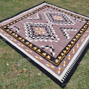 Pode incluir: Um tapete quadrado tecido com um padrão geométrico em tons de castanho, preto, branco e amarelo. O tapete apresenta um design central com formas repetidas e uma borda com uma borda preta contrastante. O tapete está colocado sobre relva verde.