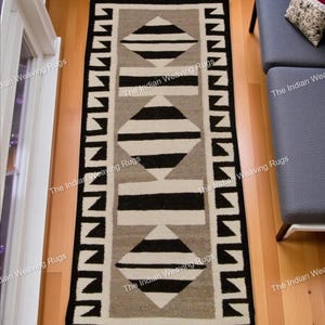 Puede incluir: Una alfombra rectangular con un diseño geométrico. La alfombra presenta un patrón central de formas de rombos negros y blancos alternos con rayas horizontales negras, sobre un fondo gris. El borde es negro con formas geométricas blancas.
